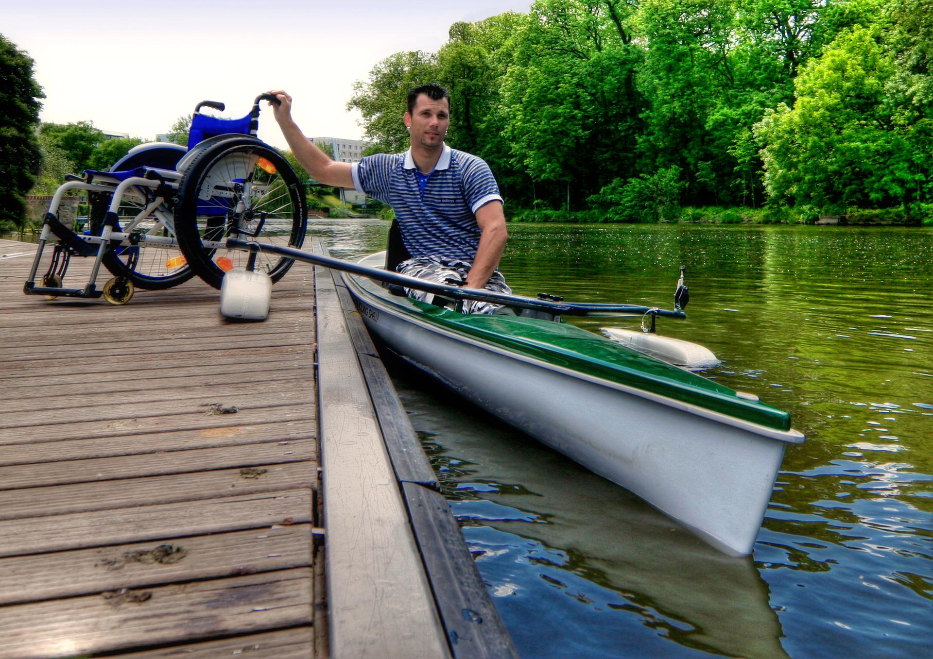 A man in a kayak holds onto the dock with one hand and his wheelchair with the other. The kayak is on a calm river surrounded by lush green trees. The dock extends beside him.