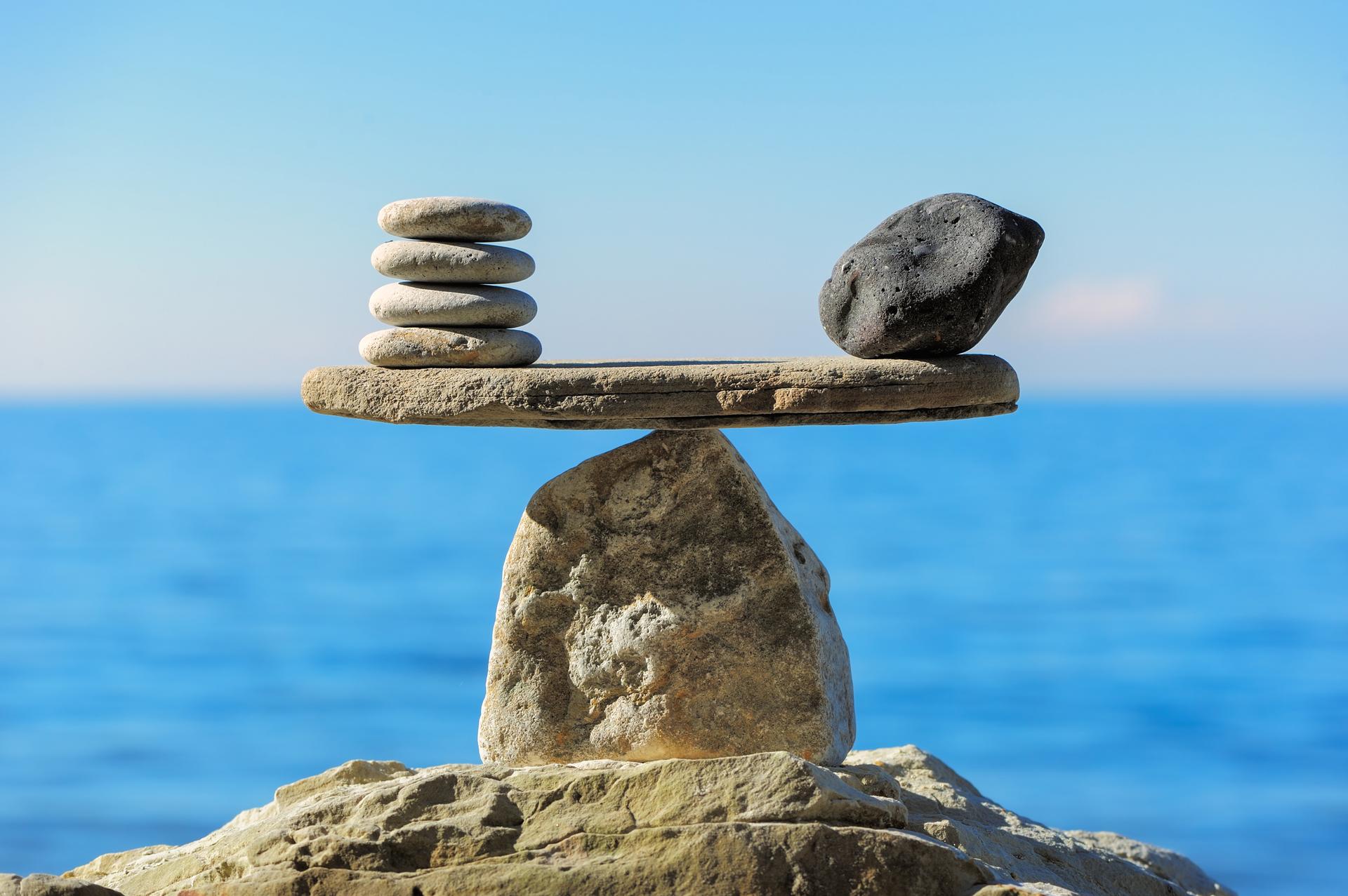 Five smooth white pebbles and one larger dark stone are balanced on a flat rock, which is supported by a pointed stone, with the sea and blue sky in the background.