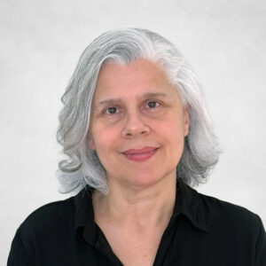 A woman with shoulder-length grey hair and a slight smile, wearing a black button-up shirt, stands in front of a plain light background.