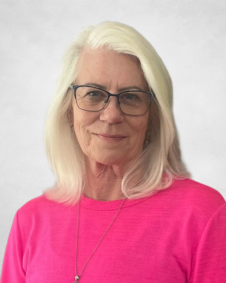 Bonnie Nybo, A woman with long white hair and glasses, wearing a bright pink top and a necklace, poses in front of a plain light grey background.