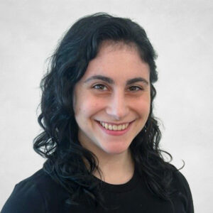 Daniela Luongo, A woman with wavy dark hair wearing a black top smiles at the camera against a plain, light grey background.