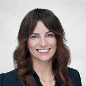Danielle Rae, A woman with wavy brown hair wearing a dark blouse smiles at the camera against a light, neutral background.