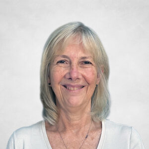 Debbie Field, Smiling older woman with straight, shoulder-length grey-blonde hair, wearing a white top and a necklace with a small pendant, standing in front of a plain light grey background.