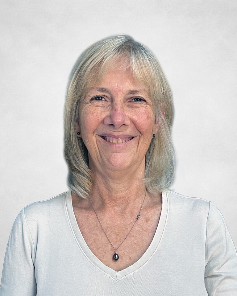 Debbie Field is a smiling older woman with straight, shoulder-length grey-blonde hair. She is wearing a white top and a necklace with a small pendant.