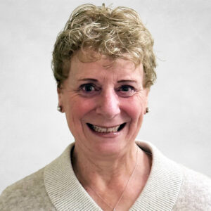 An older woman with short, curly blonde hair smiles at the camera. She wears a light beige jumper with a wide collar and a silver necklace. The background is plain and light-coloured.