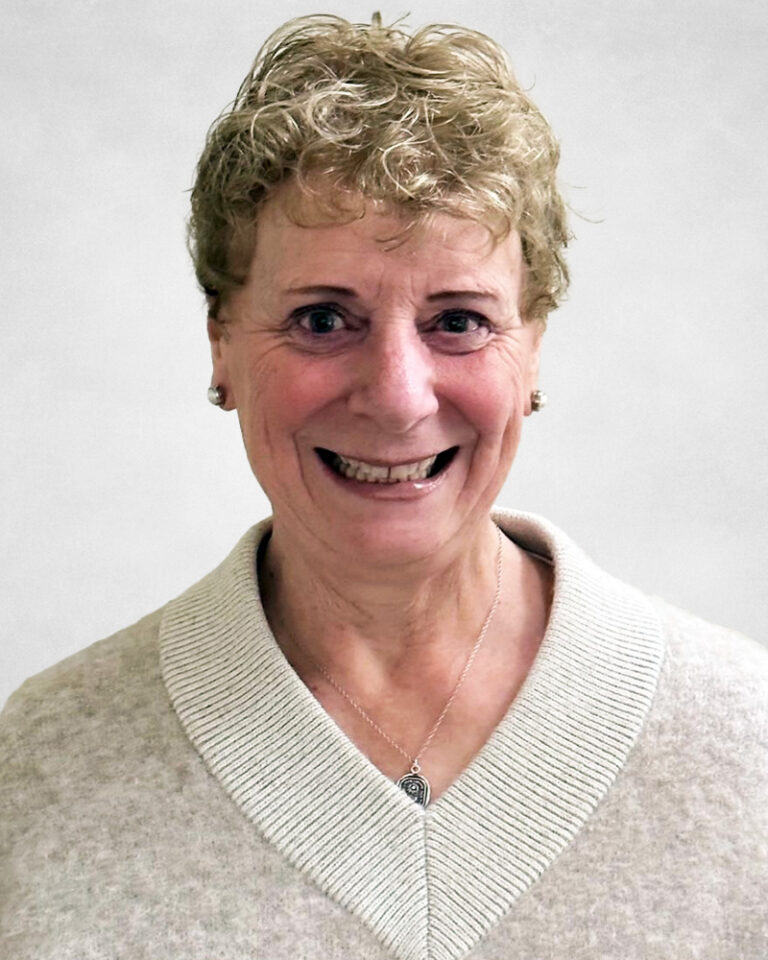 Diane Gerrard is an older woman with short, curly blonde hair. She wears a light beige jumper with a wide collar and a silver necklace.