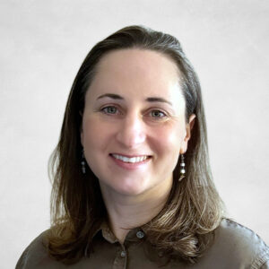 A woman with straight, shoulder-length brown hair smiles at the camera. She is wearing a brown button-up shirt and dangling earrings. The background is plain and light-coloured.