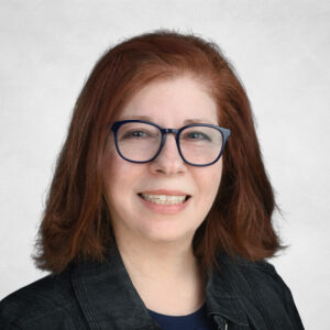 A woman with shoulder-length reddish-brown hair, wearing glasses, a dark jacket, and a navy blue shirt, smiles at the camera against a light grey background.