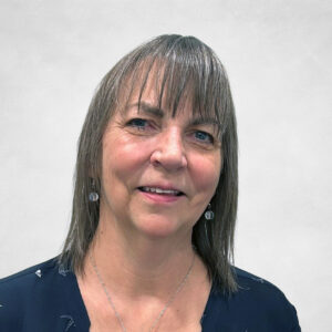 A middle-aged woman with straight, shoulder-length light brown hair and fringe smiles gently. She wears a navy blouse with a heart-shaped pendant necklace and small earrings, in front of a light grey background.