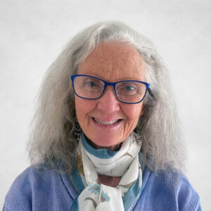 Janice Duivestein, An older woman with long grey hair and blue glasses smiles at the camera. She is wearing a lavender jumper and a white scarf with a blue floral pattern, standing against a plain light background.