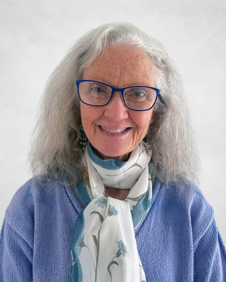 Janice Duivestein is an older woman with long grey hair and blue glasses. She is wearing a lavender sweater and a white scarf with a blue floral pattern.