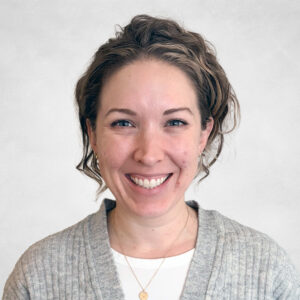 A woman with wavy, light brown hair smiles at the camera. She is wearing a grey cardigan over a white shirt and a gold necklace with a small circular pendant. The background is plain and light-coloured.