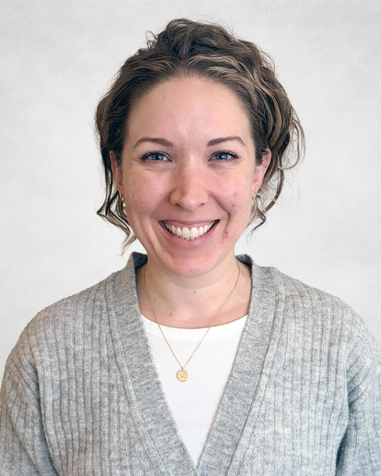 A woman with wavy, light brown hair smiles at the camera. She is wearing a grey cardigan over a white shirt and a gold necklace with a small circular pendant. The background is plain and light-coloured.