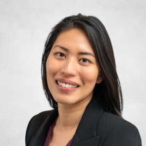A woman with long black hair, wearing a black blazer and burgundy top, smiles at the camera against a light grey background.