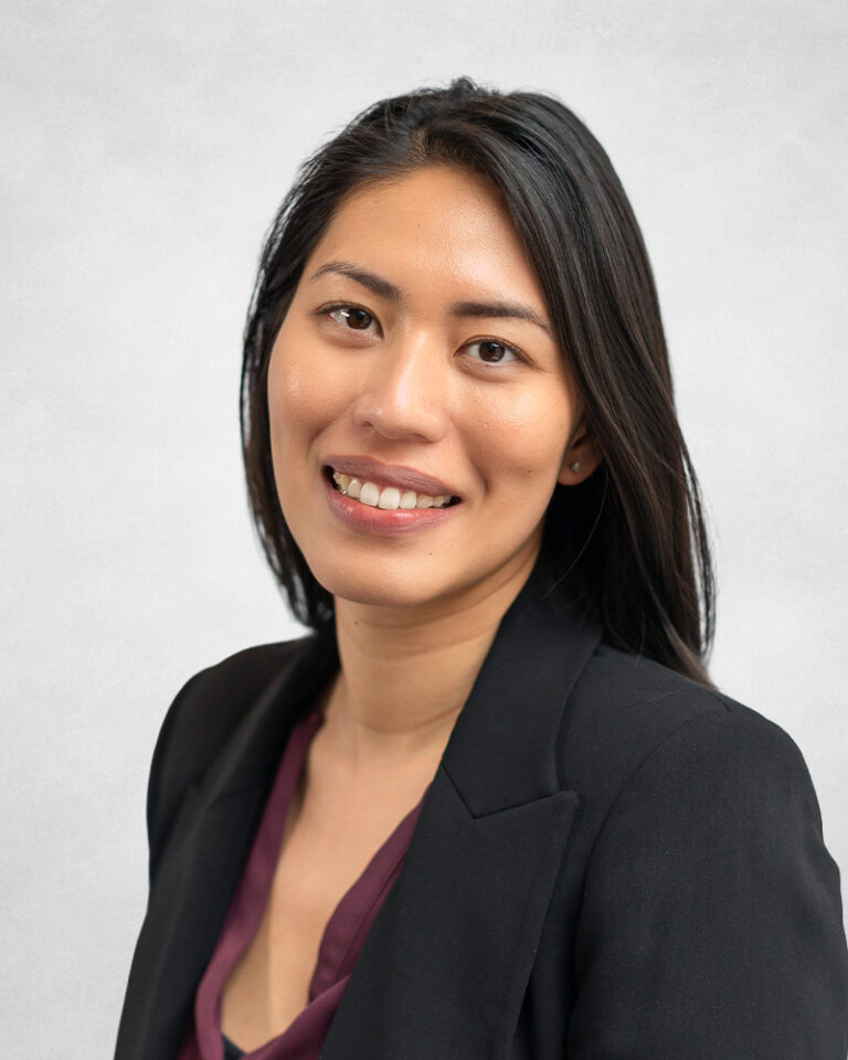 A woman with long black hair, wearing a black blazer and burgundy top, smiles at the camera against a light grey background.