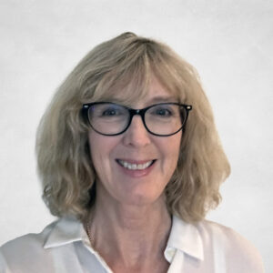 Jo Anne Chisholm, A smiling woman with shoulder-length blonde hair and glasses, wearing a white collared shirt, stands in front of a plain light background.