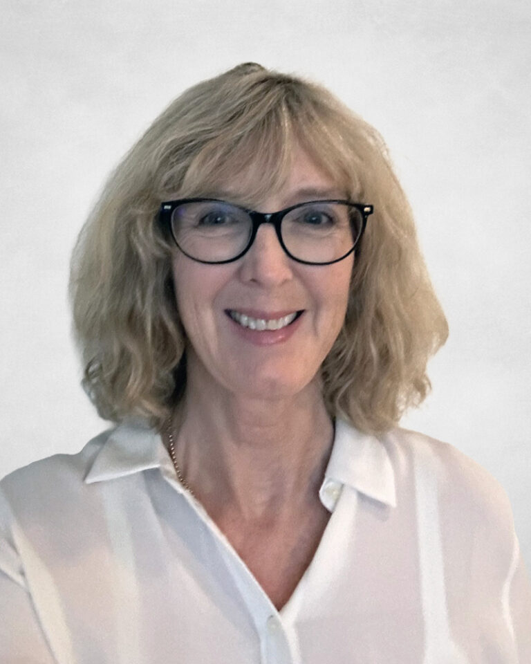 Jo Anne Chisholm, A smiling woman with shoulder-length blonde hair and glasses, wearing a white collared shirt, stands in front of a plain light background.