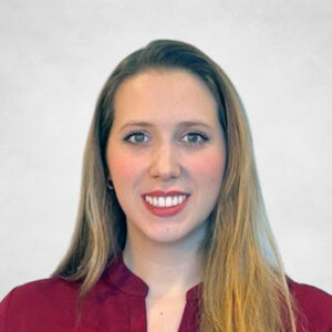 Julianna De Souza, A woman with long, straight blonde hair wearing a burgundy blouse smiles at the camera against a plain light grey background.