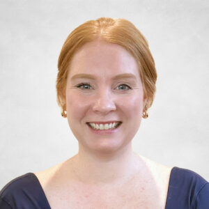 Karalyn Mayne, A woman with light skin and red hair pulled back smiles at the camera. She is wearing gold hoop earrings and a navy blue top with a scoop neckline, set against a plain light background.