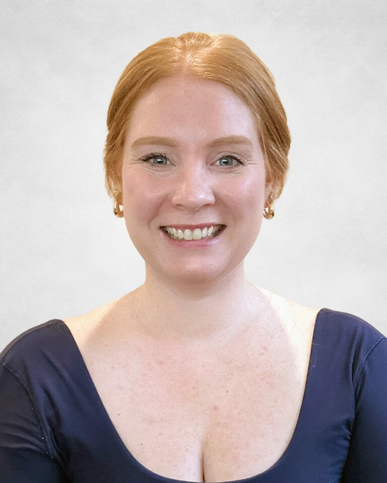 Karalyn Mayne, A woman with light skin and red hair pulled back smiles at the camera. She is wearing gold hoop earrings and a navy blue top with a scoop neckline, set against a plain light background.