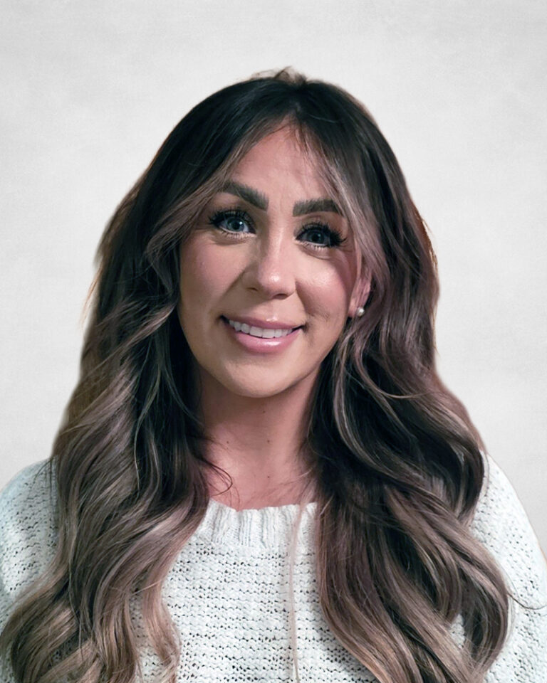 Lauren Saunders, A woman with long, wavy brown hair wearing a white knitted jumper smiles in front of a light grey background.