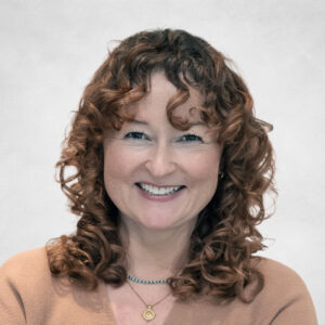 Lindsay Alford, A woman with curly brown hair and fair skin smiles confidently. She is wearing a light brown top and two necklaces, one with a gold pendant. The background is plain and light-coloured.