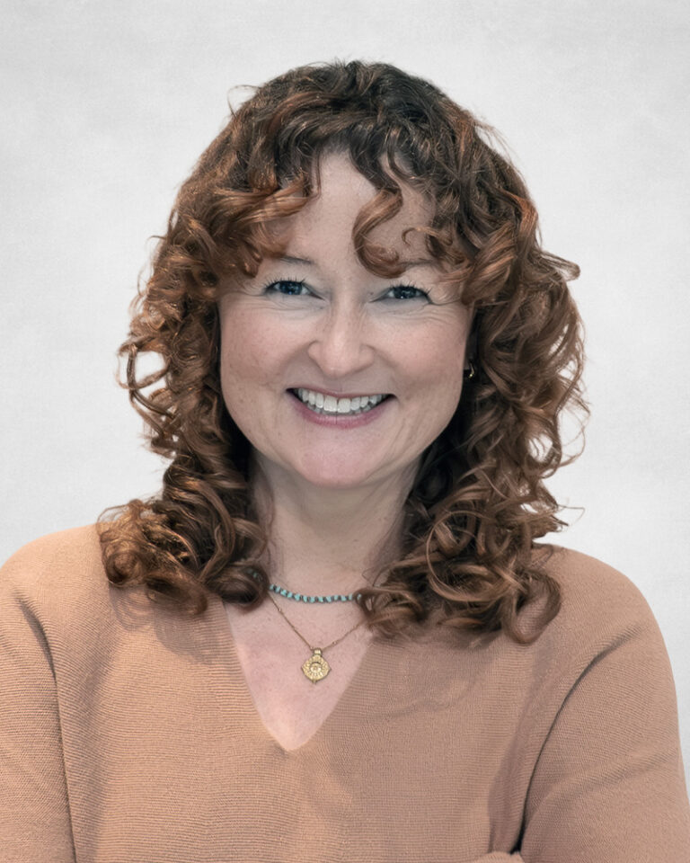 Lindsay Alford, A woman with curly brown hair and fair skin smiles confidently. She is wearing a light brown top and two necklaces, one with a gold pendant. The background is plain and light-coloured.