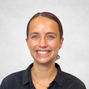 Lucy Friend, A woman with fair skin and blue eyes, smiling, wearing a black button-up shirt and a gold necklace. Her hair is tied back, and she is standing in front of a plain, light-coloured background.