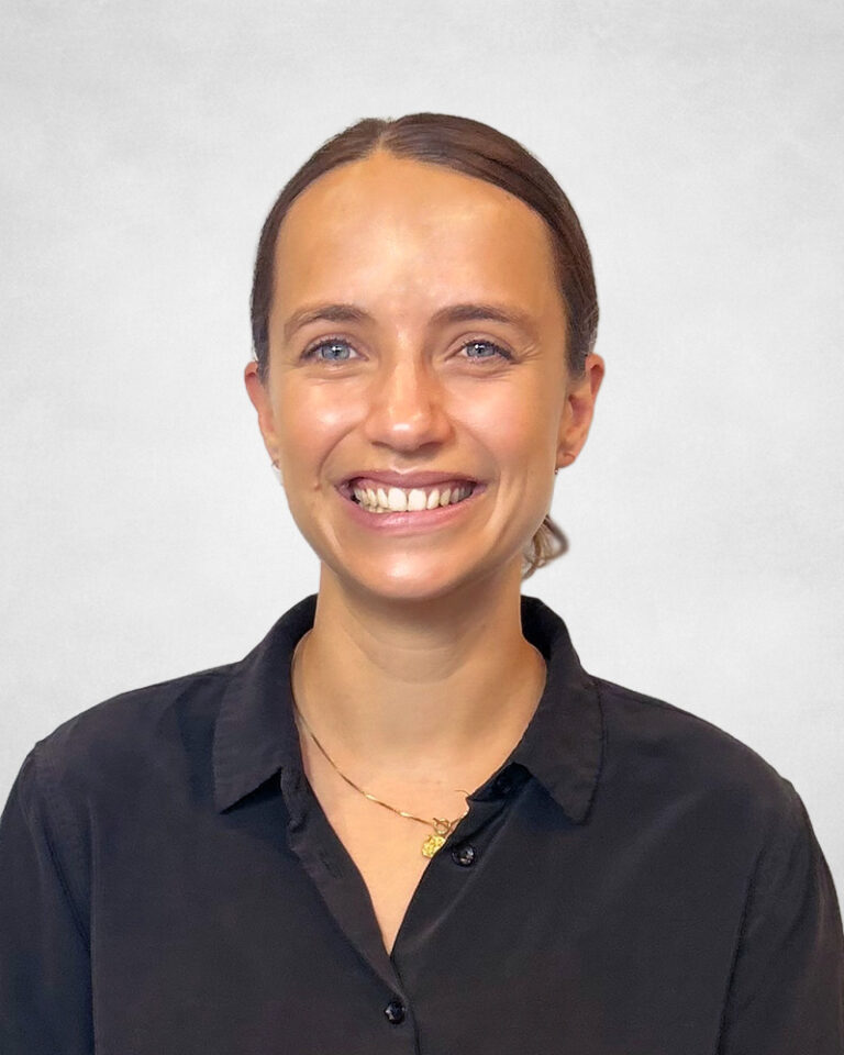Lucy Friend, A woman with fair skin and blue eyes, smiling, wearing a black button-up shirt and a gold necklace. Her hair is tied back, and she is standing in front of a plain, light-coloured background.