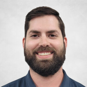 Mike Askew, A man with short dark hair and a full beard is smiling at the camera. He is wearing a dark blue collared shirt and is posed against a light grey background.