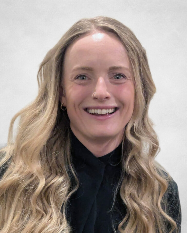Montana Bahen, A smiling woman with long, wavy blonde hair, wearing a black top, poses in front of a light grey background.