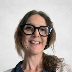Najade Minns, A woman with wavy brown hair, wearing large black glasses, a navy blouse, a light jumper, and a gold necklace, smiling in front of a plain light background.