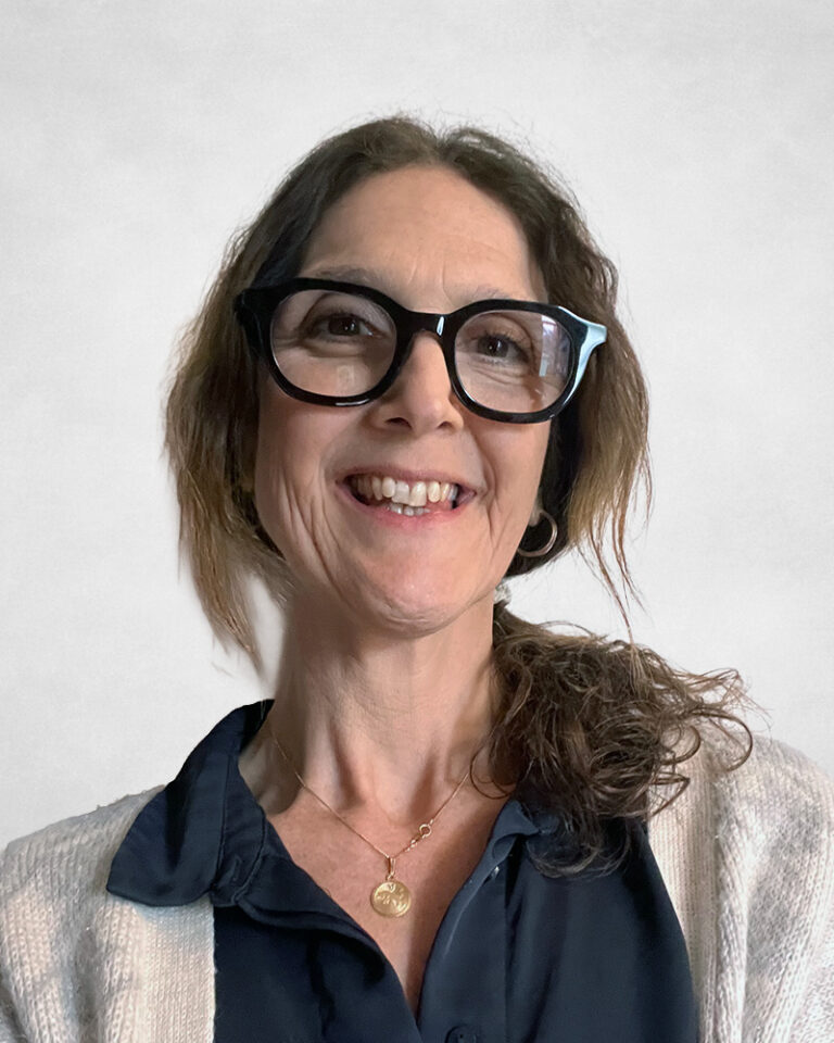 Najade Minns, A woman with wavy brown hair, wearing large black glasses, a navy blouse, a light jumper, and a gold necklace, smiling in front of a plain light background.