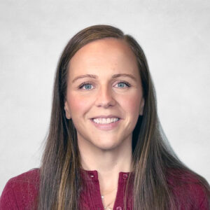Nicole Armstrong, A woman with long brown hair, wearing a maroon knitted jumper, smiles at the camera against a light grey background.