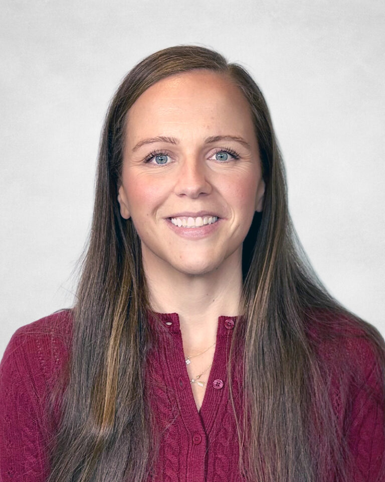 Nicole Armstrong, A woman with long brown hair, wearing a maroon knitted jumper, smiles at the camera against a light grey background.