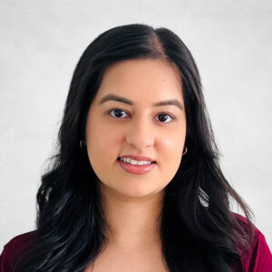 Preety Sian, A woman with long, wavy black hair and hoop earrings is smiling slightly. She is wearing a burgundy top and is posed against a light grey background.