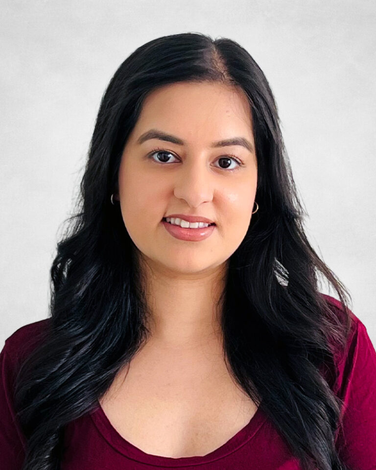 Preety Sian, A woman with long, wavy black hair and hoop earrings is smiling slightly. She is wearing a burgundy top and is posed against a light grey background.