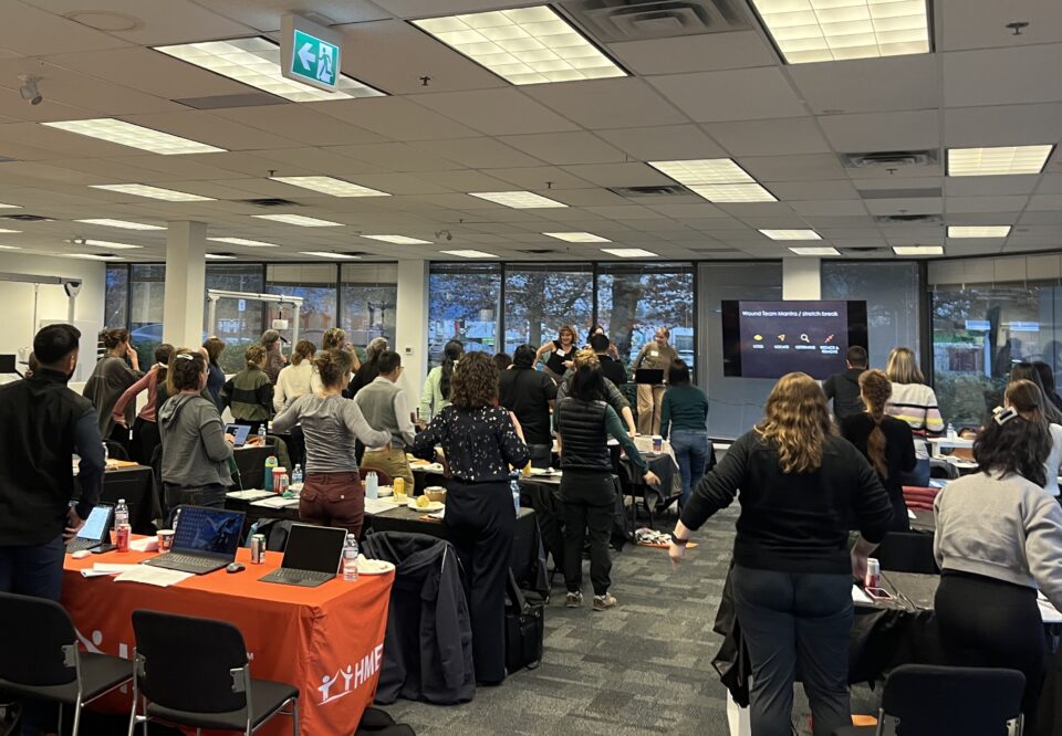 A group of people stands and stretches together in a conference room during a workshop or seminar, facing a presentation screen. Tables with laptops, drinks, and materials are visible throughout the room.