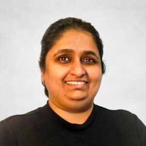 Samanthan Singh, A person with dark hair pulled back, wearing a black top, smiles whilst looking slightly to the right. The background is plain and light-coloured.