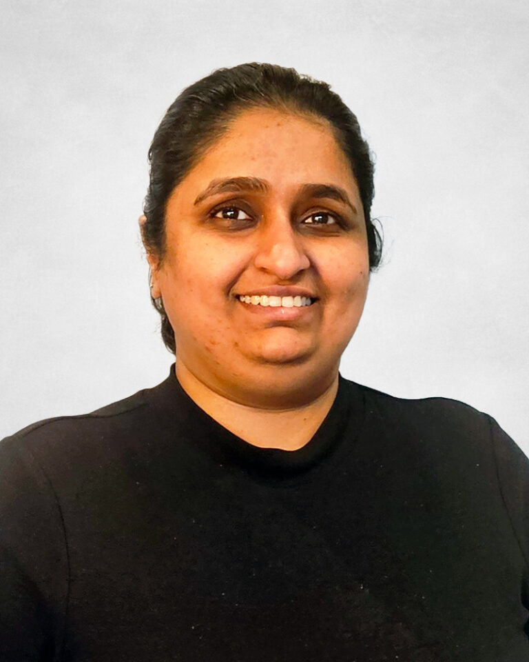 Samanthan Singh, A person with dark hair pulled back, wearing a black top, smiles whilst looking slightly to the right. The background is plain and light-coloured.