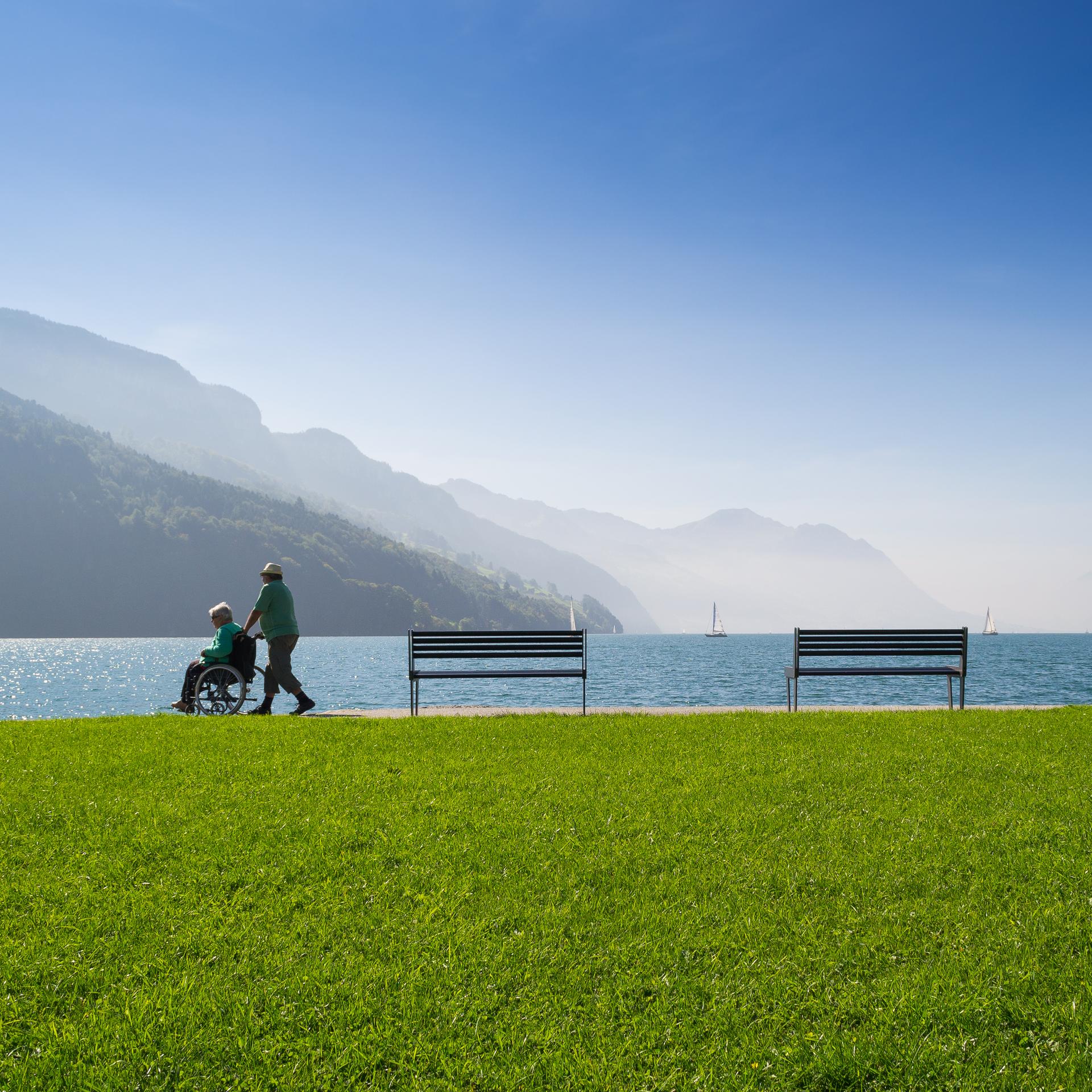 A person pushes another in a wheelchair along a lakeside path with benches, green grass, calm water, and mountains under a clear blue sky in the background.