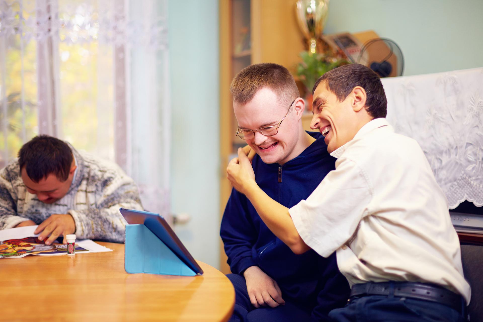 Two men with Down's syndrome sit at a table, smiling and laughing together whilst looking at a tablet, as another man on the left draws with crayons, partially hiding his face.