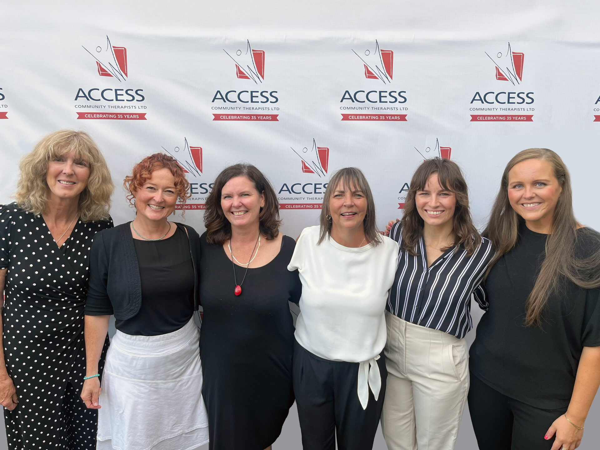 Six women stand smiling together in front of a white ACCESS Community Therapists LTD backdrop, celebrating its 30th year, dressed in black and white outfits.