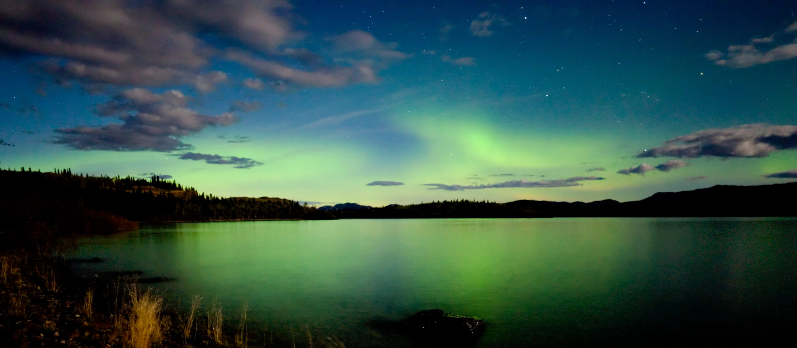 Northern lights illuminate the night sky above a calm lake, reflecting green and blue hues. Silhouetted hills and scattered clouds frame the tranquil, starry scene.