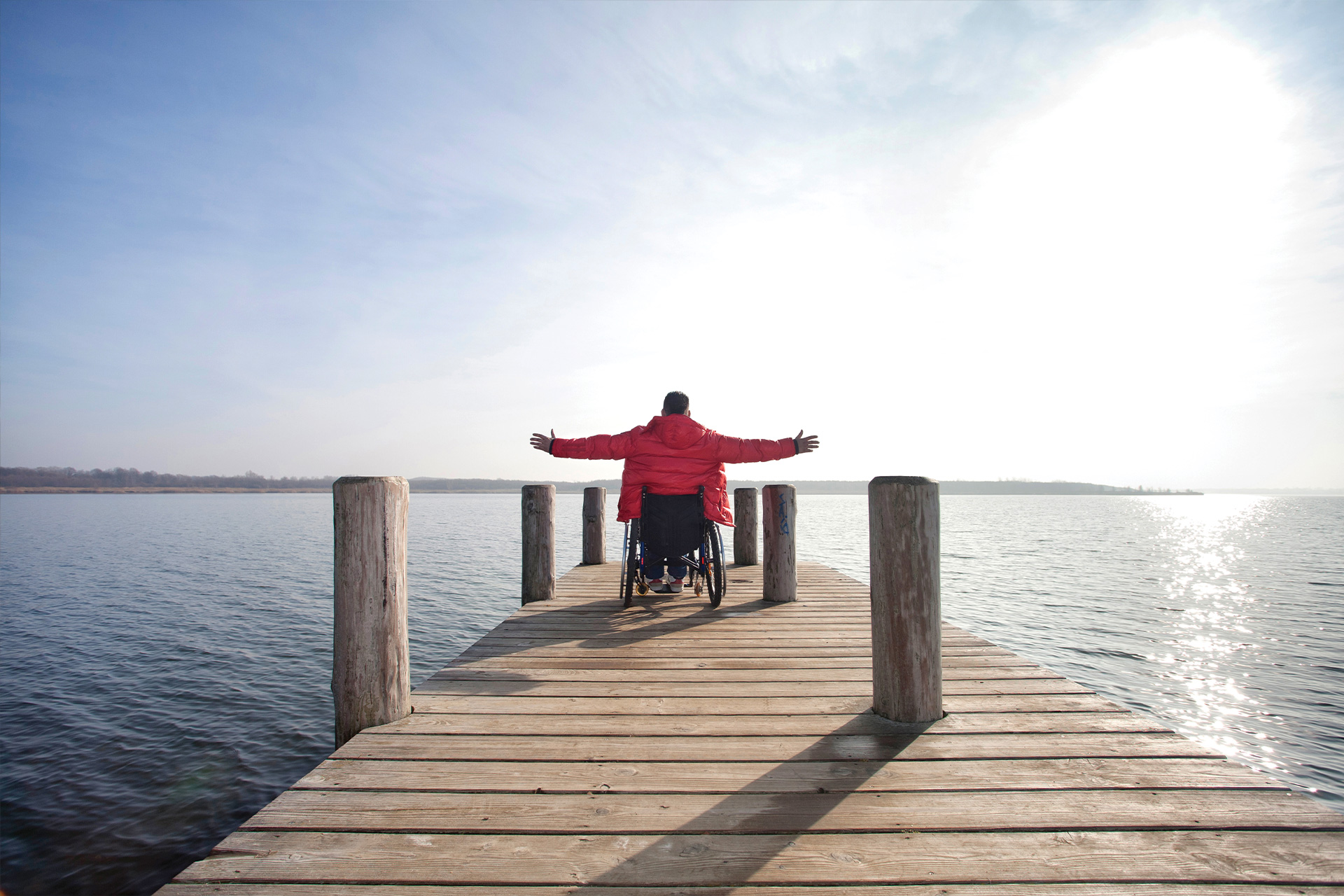 A person in a wheelchair, wearing a red coat, sits at the end of a wooden jetty with arms outstretched, facing a calm lake under a bright, partly cloudy sky.