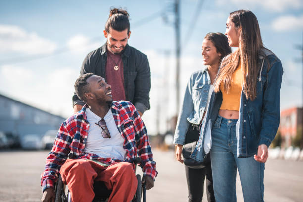 Four friends walk together outside on a sunny day. One man is in a wheelchair, smiling at his mates; another friend is pushing the wheelchair. Everyone appears happy and engaged in conversation.
