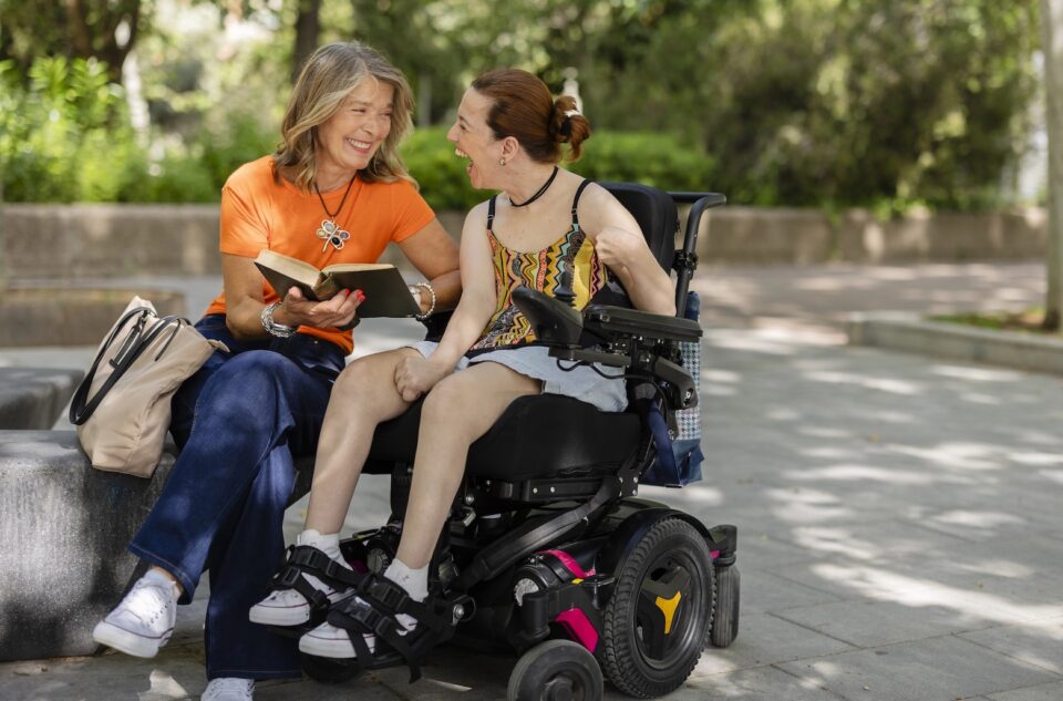 Two women sit outdoors, smiling and laughing together. One woman holds a book and sits on a bench, while the other, wearing a summer dress, sits in an electric wheelchair. Trees and greenery are visible in the background.