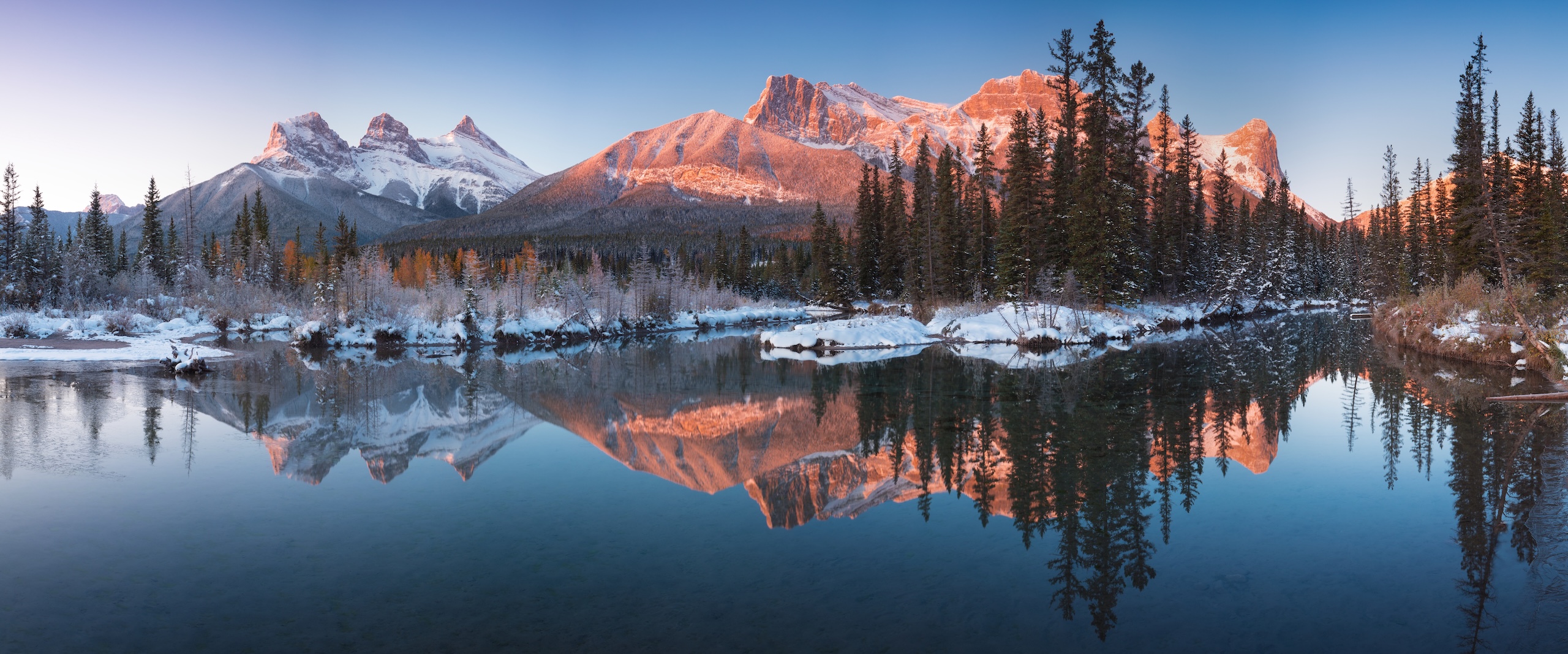 Snow-capped mountain peaks and pine trees are reflected in a calm, clear lake at sunrise. The sky is clear, and the landscape is dusted with snow, creating a serene and picturesque winter scene.
