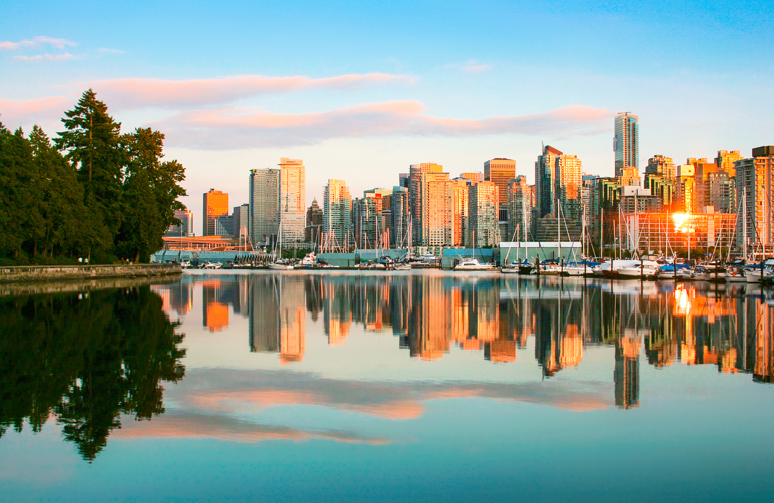 A city skyline with tall buildings is reflected in calm water at sunset, with boats moored at a marina and trees visible on the left side of the image.