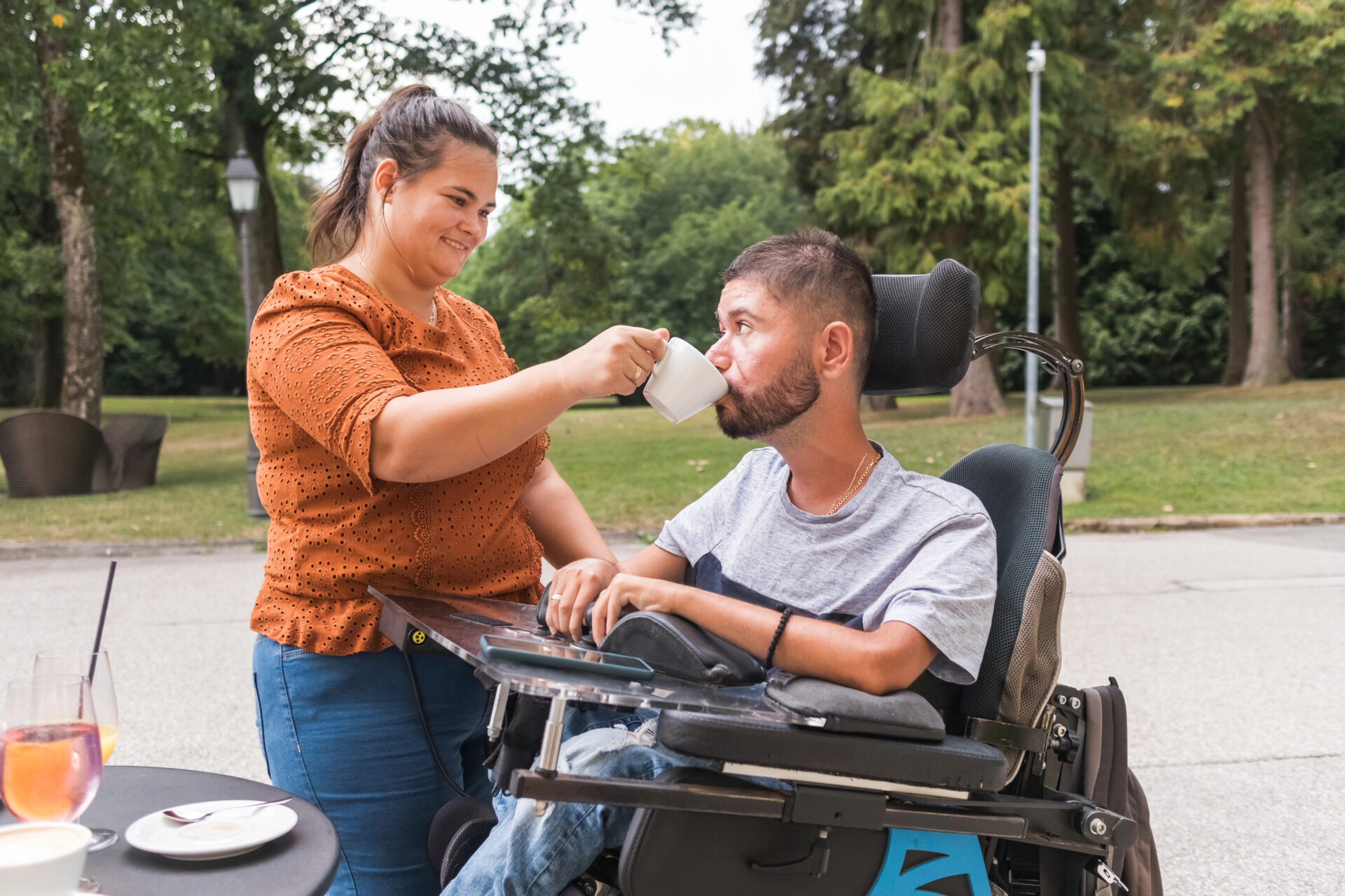 A woman helps a man in a wheelchair drink from a mug outdoors in a park. The man looks at her whilst she holds the mug to his mouth. They both appear calm and engaged with each other.